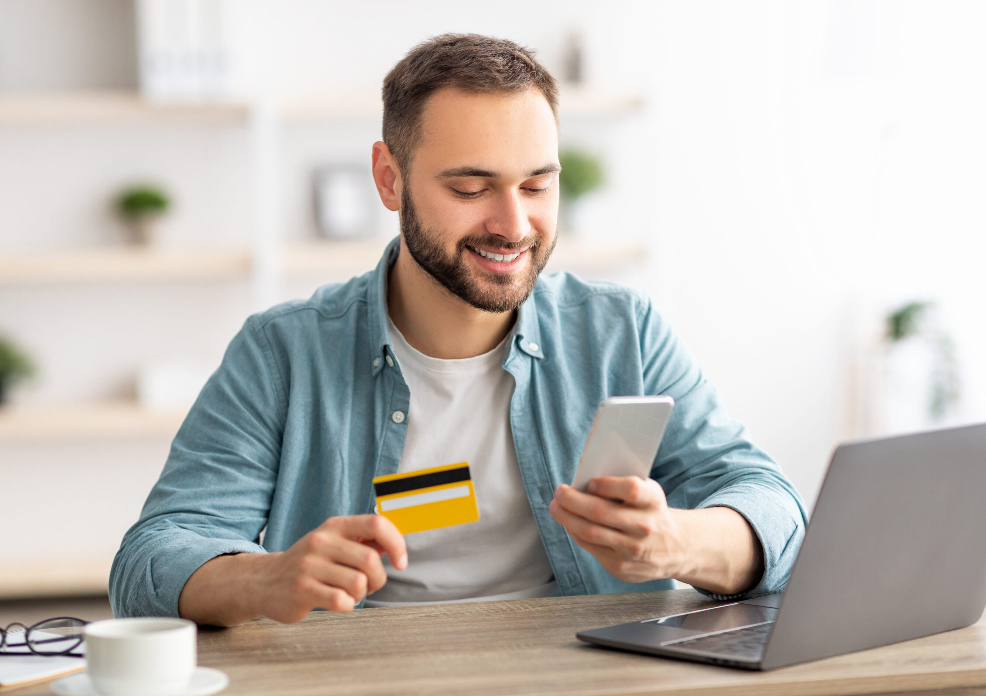 Happy Caucasian man buying things online, using smartphone, laptop and credit card, enjoying shopping in internet. Positive young guy purchasing goods on web, making remote payment